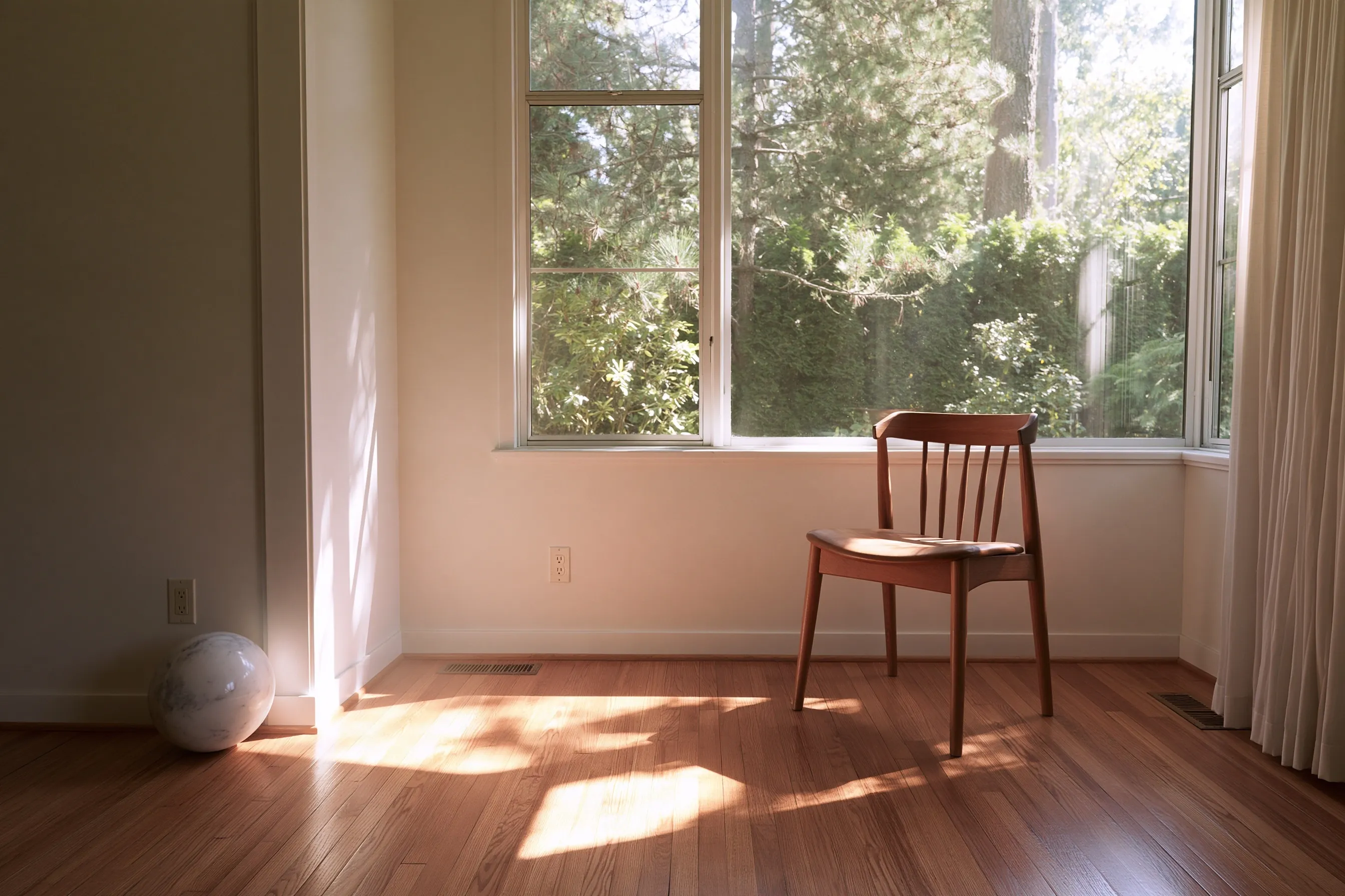 Empty room with a hardwood floor, a wooden chair by a large window onto trees, and a round ball resting on the floor against the wall at the left